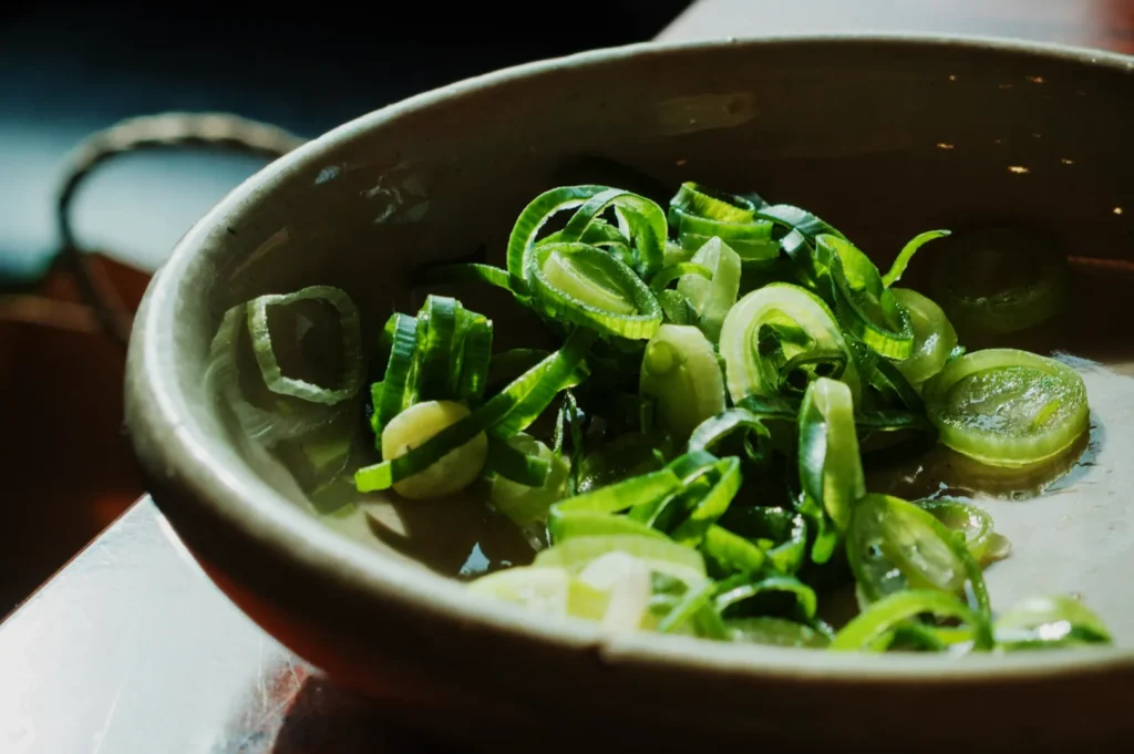 A ceramic bowl filled with freshly sliced green onions sits on a sunny countertop. The vibrant greens are highlighted by the soft natural light.