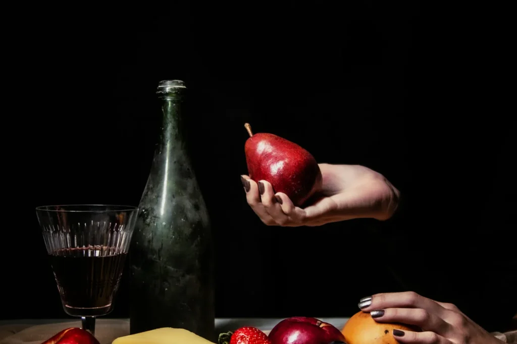 A hand holds a red pear above a table with fruit, cheese, and a dark bottle. A glass of red wine is nearby. The mood is artistic and dramatic.