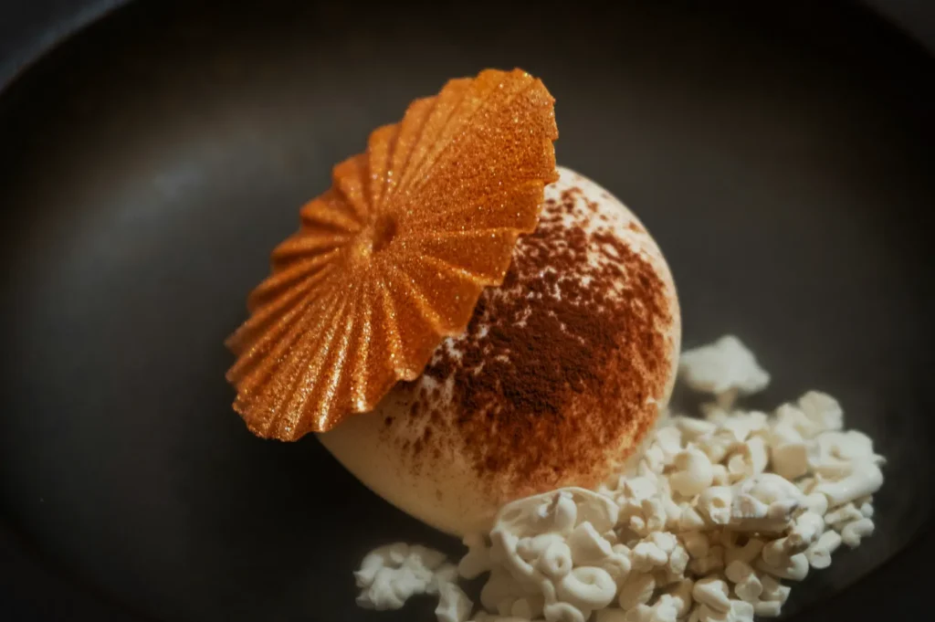 A dessert featuring a brown-dusted dome-shaped mousse topped with a sparkly golden disc, accompanied by a cluster of white popcorn, set against a dark plate.