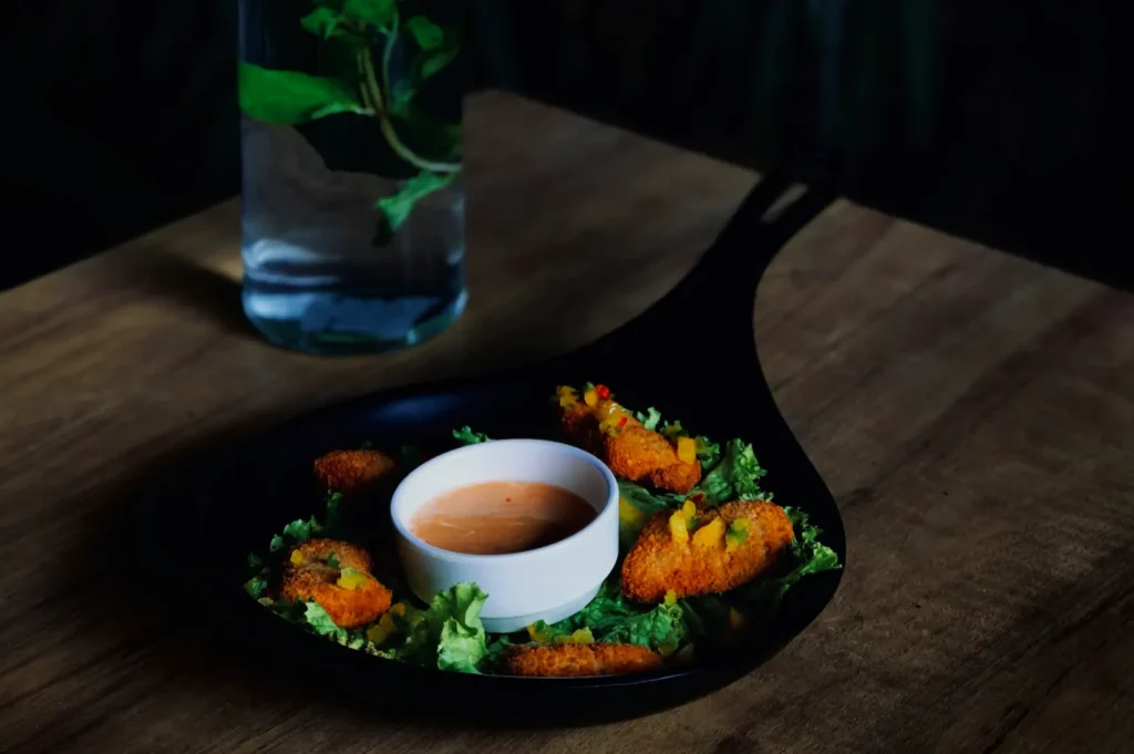 A black skillet holds crispy appetizers on a bed of lettuce with a cup of dipping sauce in the center. A glass of water with mint accompanies the dish.