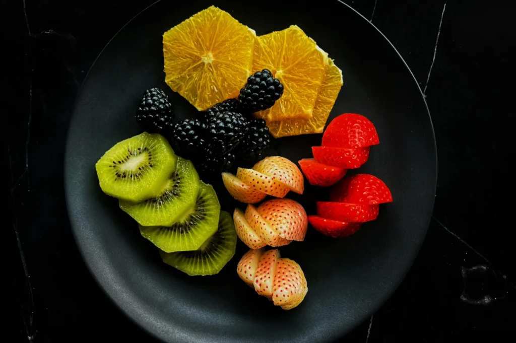 A black plate displays a colorful fruit arrangement: sliced oranges, blackberries, vibrant kiwis, and red and white strawberries, on a dark background.