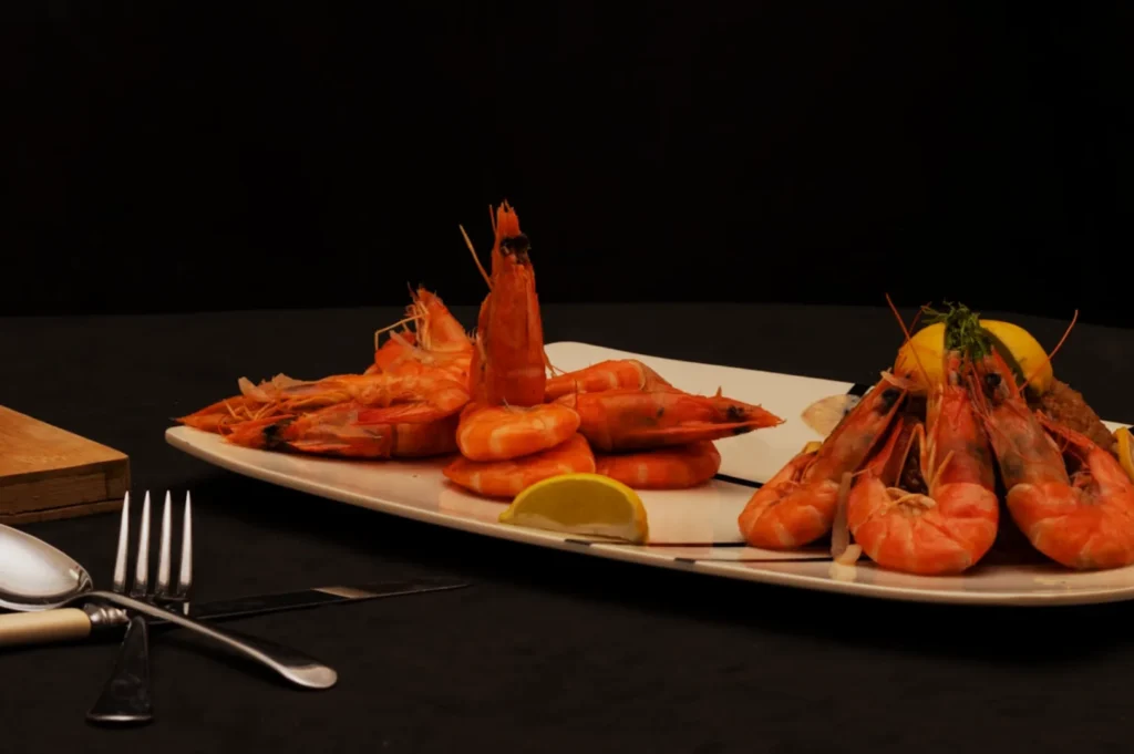 A platter of cooked prawns presented decoratively, surrounded by lemon wedges. Cutlery is placed nearby on a dark tablecloth, creating an elegant dining setting.