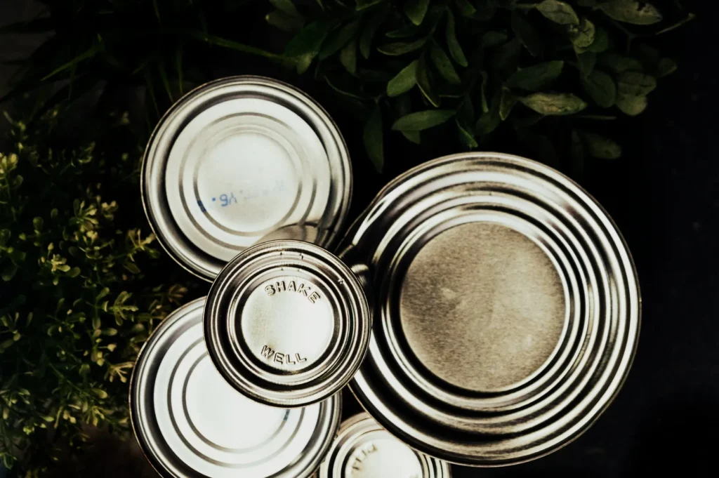 Five stacked tin cans with embossed text, surrounded by lush green foliage. The image conveys a rustic, natural feel.