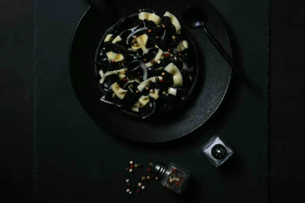 A black plate of monochrome pasta with mixed peppercorns sits on a dark slate surface. Nearby, a black spoon and pepper grinder enhance the moody atmosphere.
