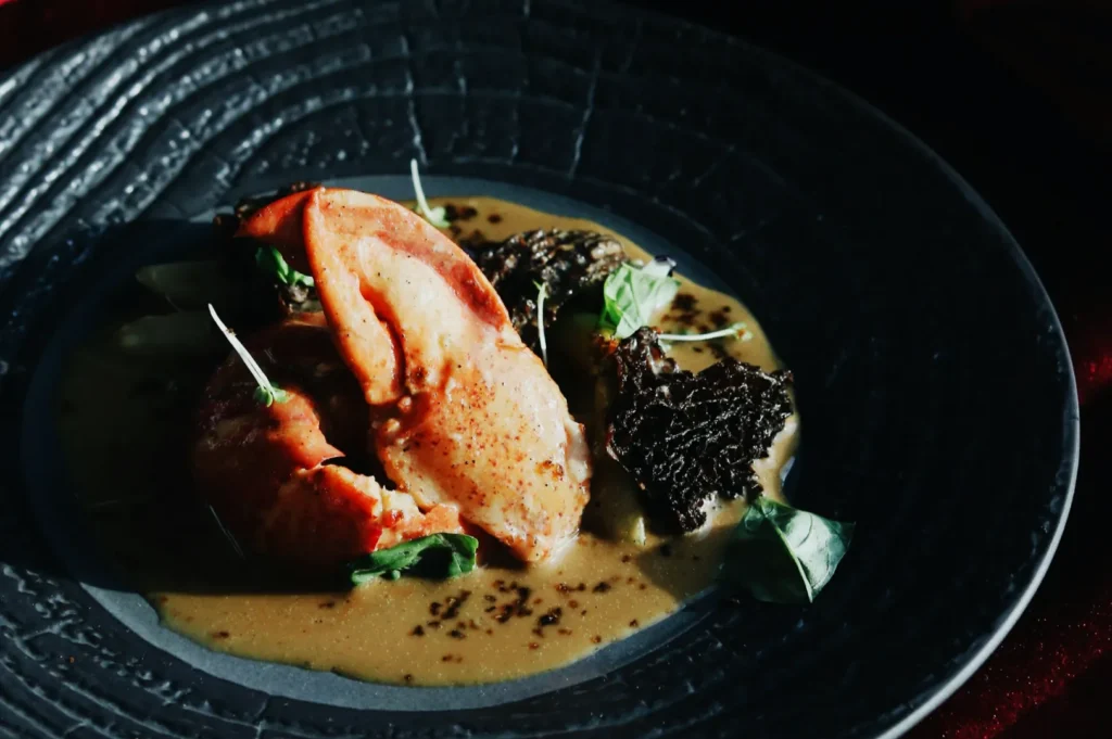 Elegant dish of lobster claw and dark mushrooms in rich brown sauce, garnished with microgreens in a textured black bowl; sophisticated and savory.