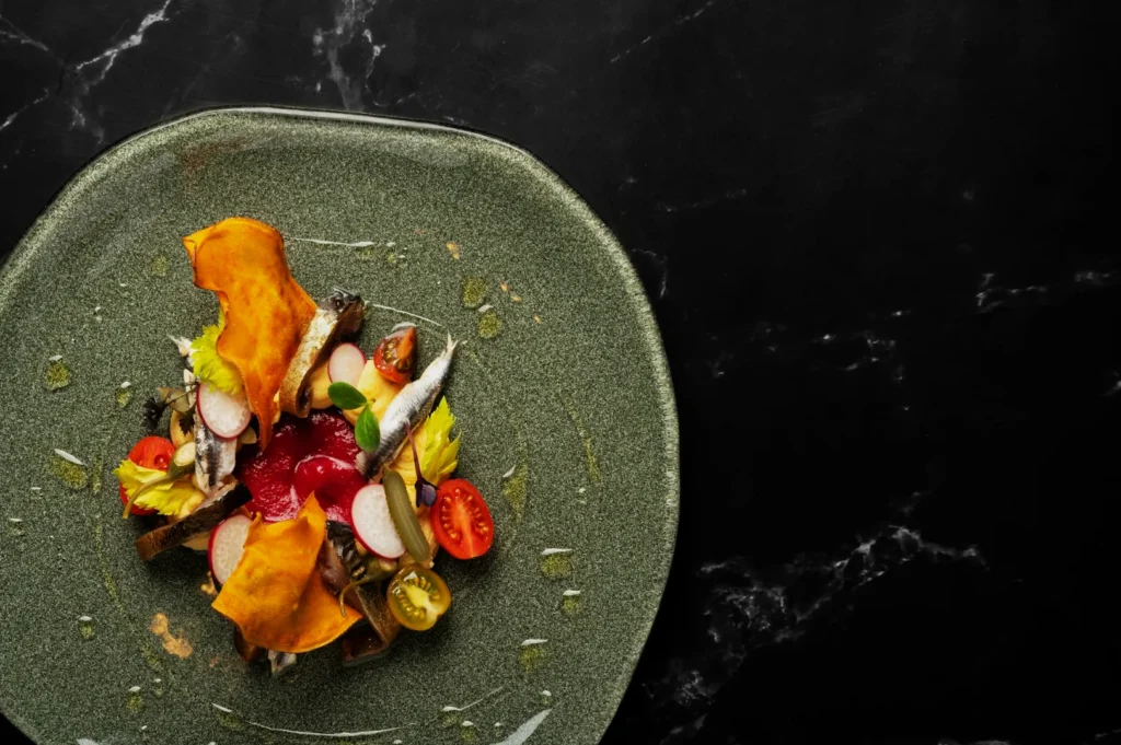 A gourmet dish is elegantly presented on a textured green plate with vibrant red beets, radish slices, small fish, cherry tomatoes, and a yellow garnish, set against a dark marble surface.