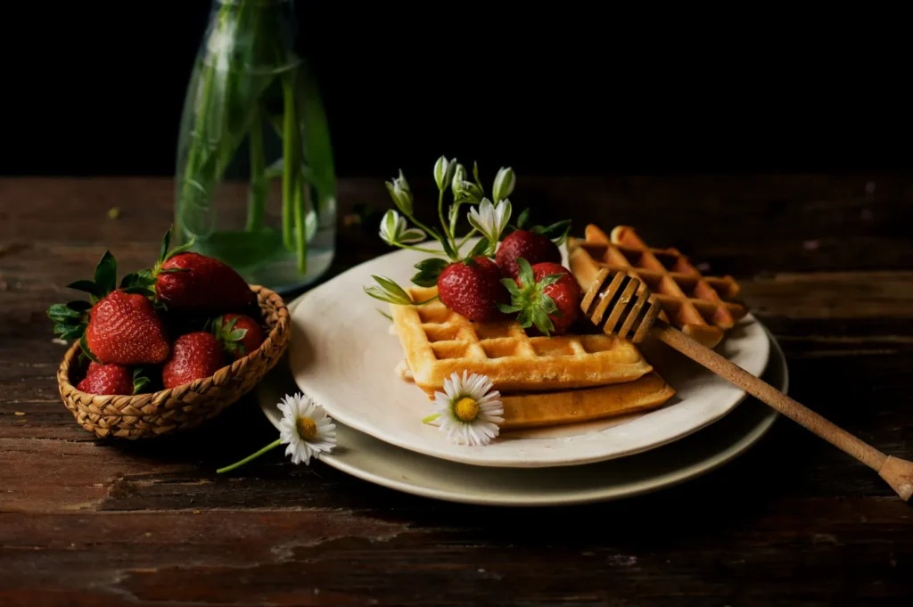 A plate of golden waffles topped with fresh strawberries and white flowers is on a wooden table. A honey dipper and small basket of strawberries are nearby.