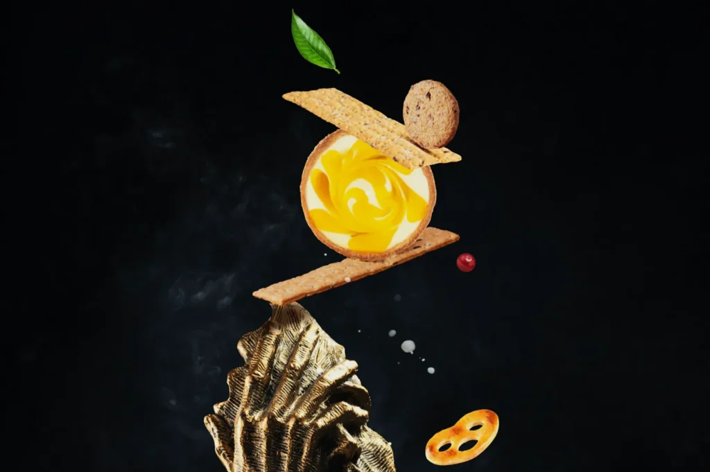A whimsical food sculpture balancing on a wooden base. It features layered crackers, a yellow cream spiral, a cookie, leaf, and floating berries on a dark background.