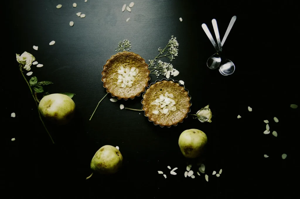 Two almond-topped tarts on a dark surface with scattered flower petals, surrounded by three pears, a white rose, and three silver spoons.