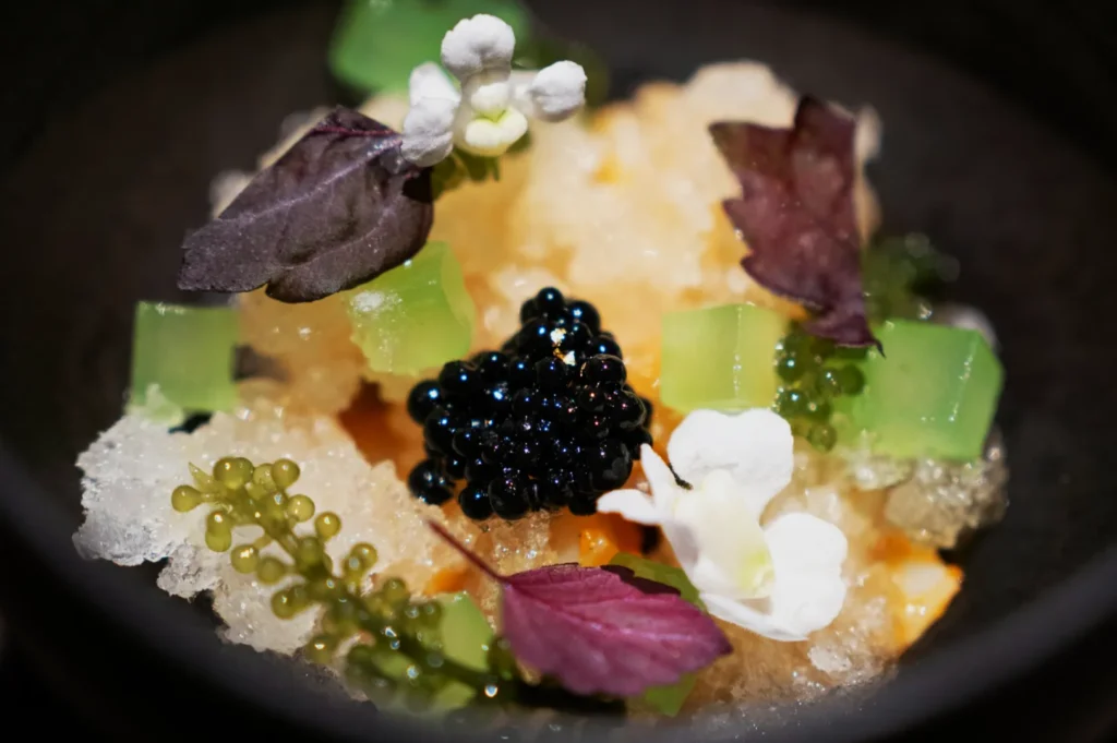 A gourmet dish featuring glistening black caviar, translucent green jelly cubes, delicate white flowers, and purple leaves on a bed of crushed ice.