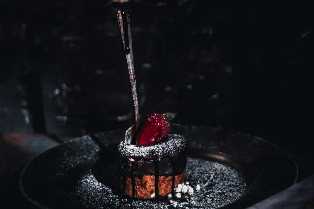 A decadent dessert on a dark plate features a chocolate-covered pastry topped with powdered sugar, a strawberry, and a fork. The mood is indulgent and elegant.