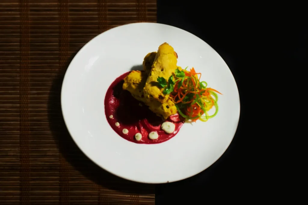 A white plate with yellow, spiced rolls on a red sauce, garnished with a swirl of carrot and green garnish. Background has a split bamboo and black design.
