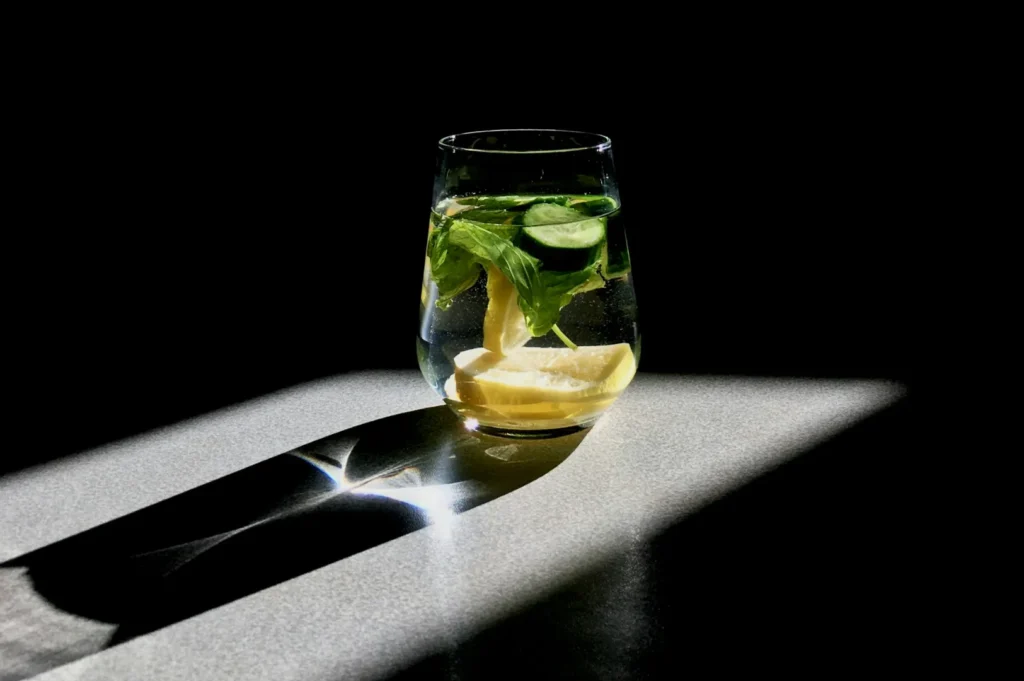 A glass of infused water with lemon, cucumber, and mint sits in sunlight on a dark surface, casting a dramatic shadow and creating a serene feel.