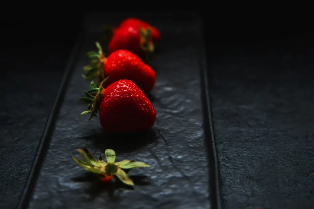 Four ripe strawberries with green tops are lined up on a dark, textured rectangular tray against a black background, conveying freshness and elegance.