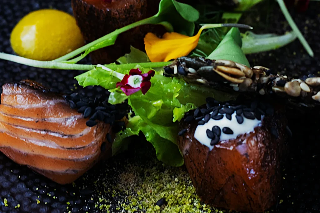 Gourmet dish with smoked salmon, topped with black sesame seeds, served with fresh greens, yellow sauce, and a seeded cracker on a textured plate.