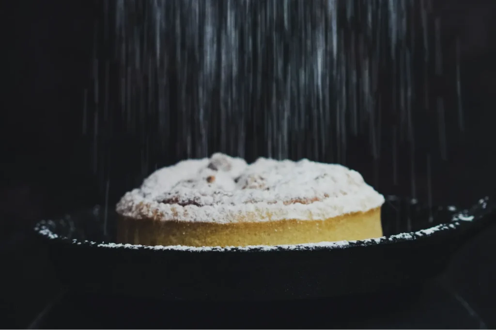 A freshly baked pie in a black pan is being dusted with powdered sugar, falling like gentle snow. The dark background enhances the cozy, rustic feel.