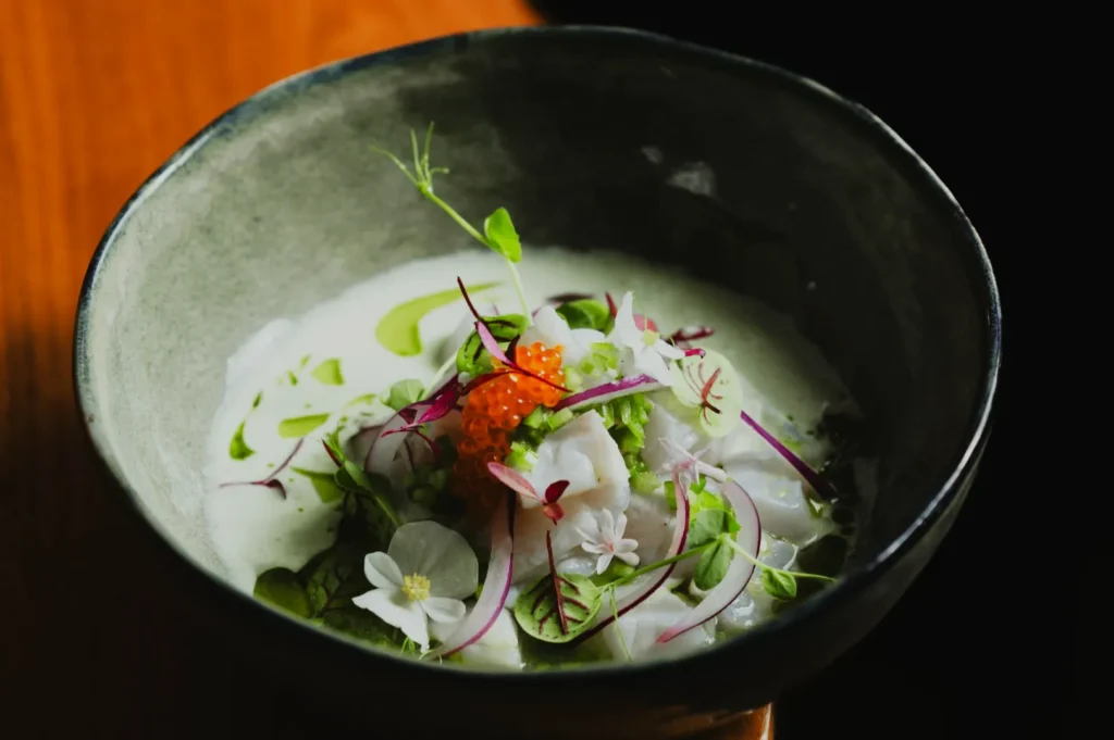 A ceramic bowl holds a gourmet dish featuring ceviche with vibrant garnishes, like orange roe, radish slices, and edible flowers, set on a wooden table.