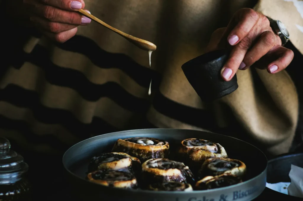 Person drizzling icing over freshly baked cinnamon rolls in a round tray. They wear a striped sweater, creating a cozy, warm atmosphere.
