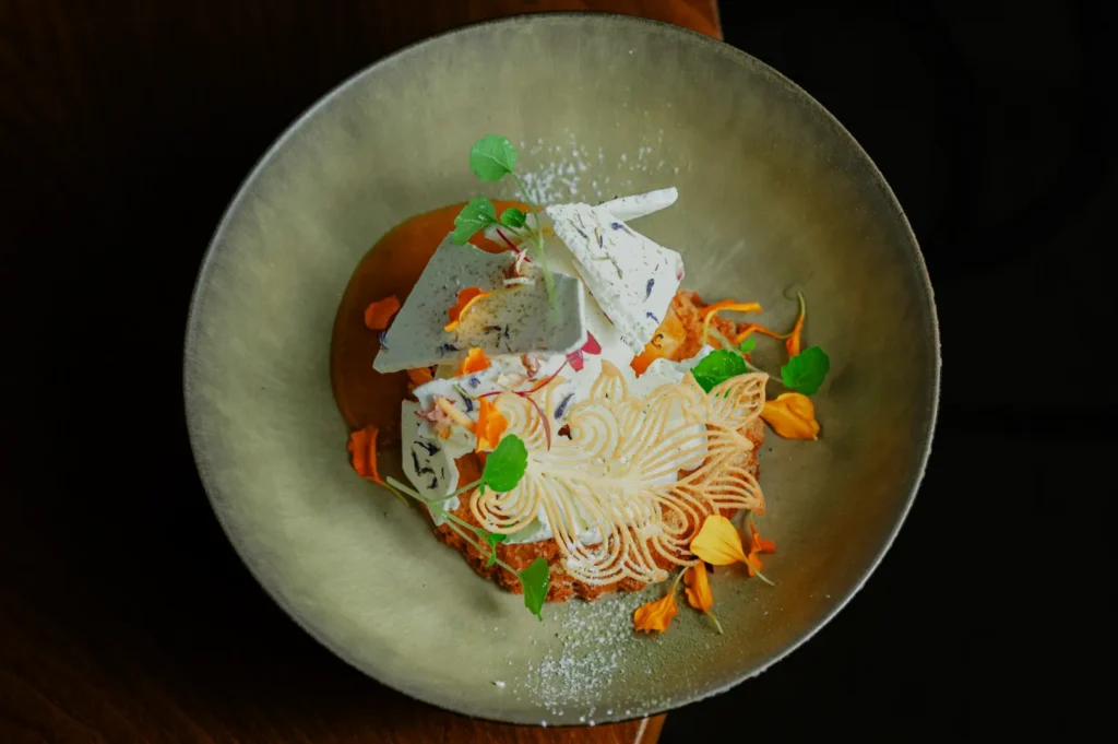 A gourmet dessert plated in a round dish, featuring whipped cream swirls, white chocolate shards, vibrant flower petals, and green leaves, creating an elegant and artistic presentation.