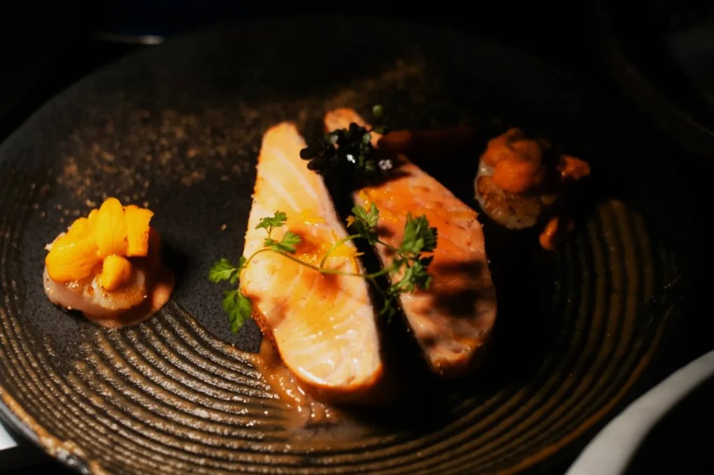 Two slices of cooked salmon garnished with herbs and surrounded by sea urchin roe on a dark textured plate, creating an elegant gourmet presentation.