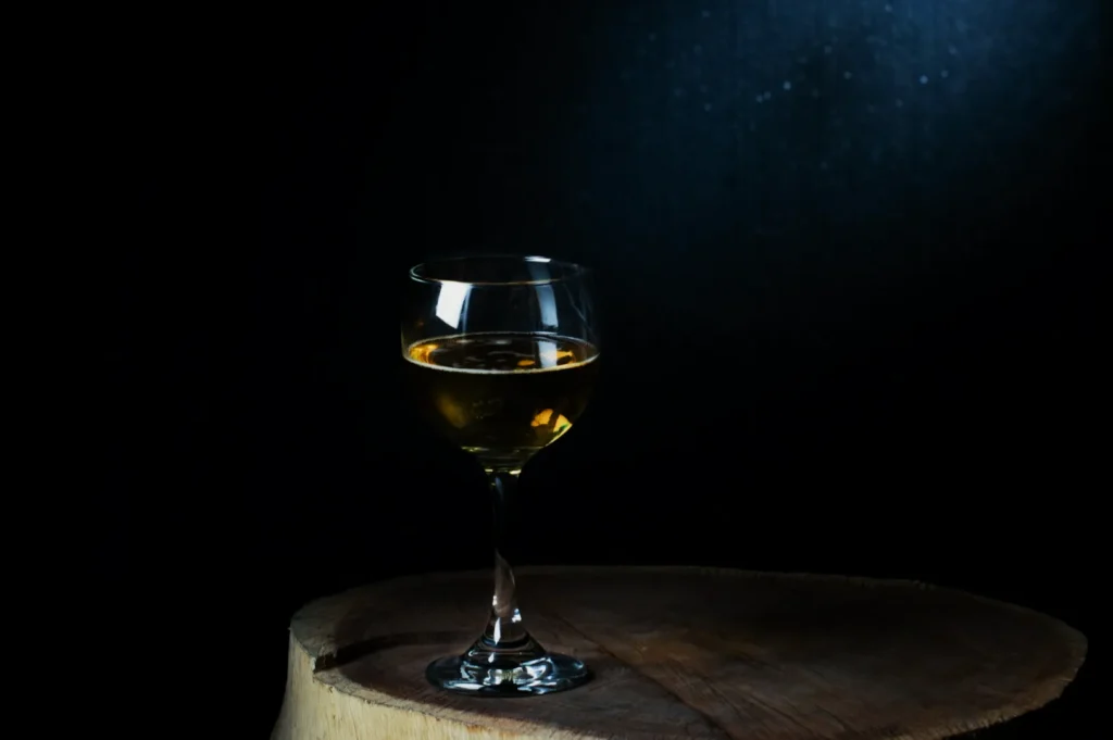 A wine glass filled with amber liquid sits on a wooden surface against a dark background. The scene has a minimalist and elegant tone.