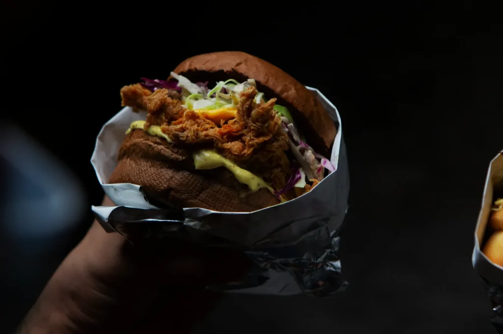 A hand holds a fried chicken sandwich wrapped in foil. The sandwich includes fried chicken, coleslaw, and sauce, with a dark, moody background.