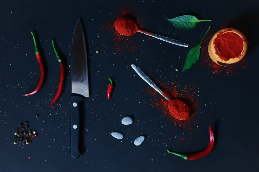 A dark surface displays scattered spices with three red chili peppers, a black-handled knife, and two spoons of red powder, conveying a bold culinary theme.