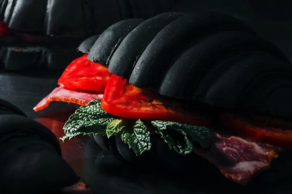 A gourmet sandwich with a black croissant bun, filled with fresh tomatoes, greens, and slices of cured meat, on a dark surface for a dramatic look.