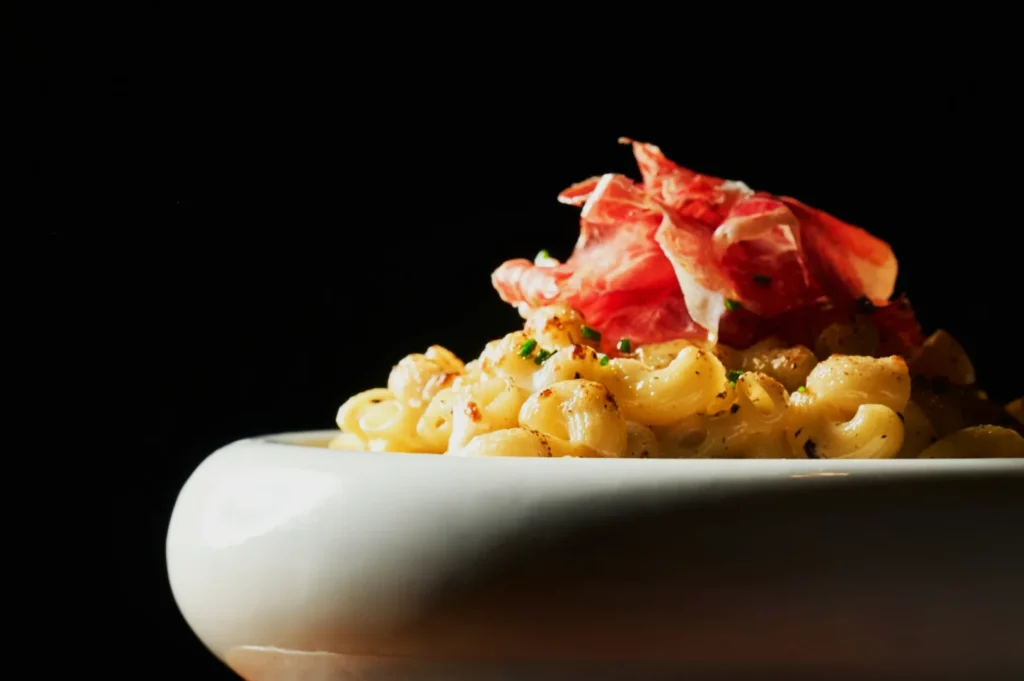 A white bowl of creamy macaroni topped with thin slices of prosciutto against a dark background. The pasta is garnished with herbs, creating a cozy, gourmet vibe.