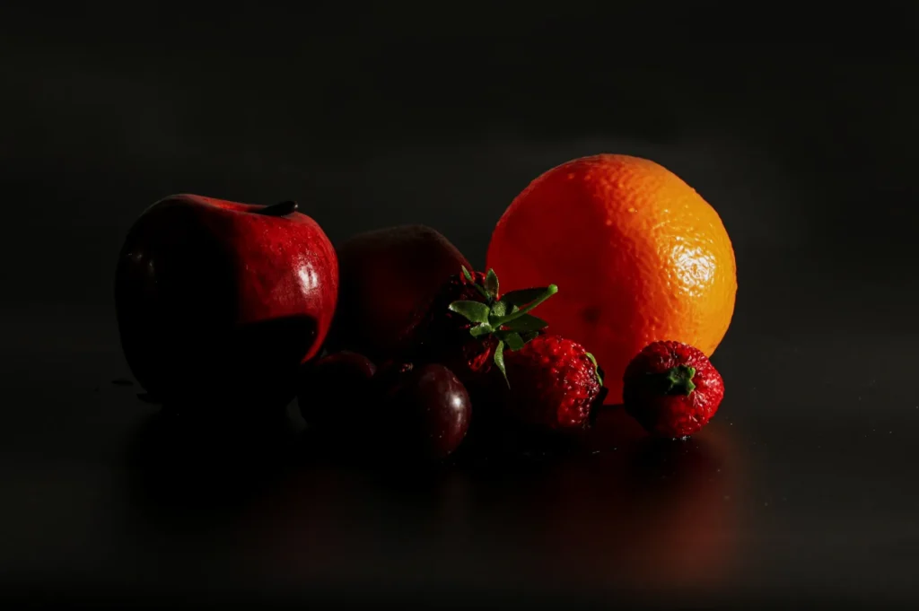 Assortment of fruits including an apple, orange, strawberries, and grapes set against a dark, moody background, highlighting vivid natural colors.