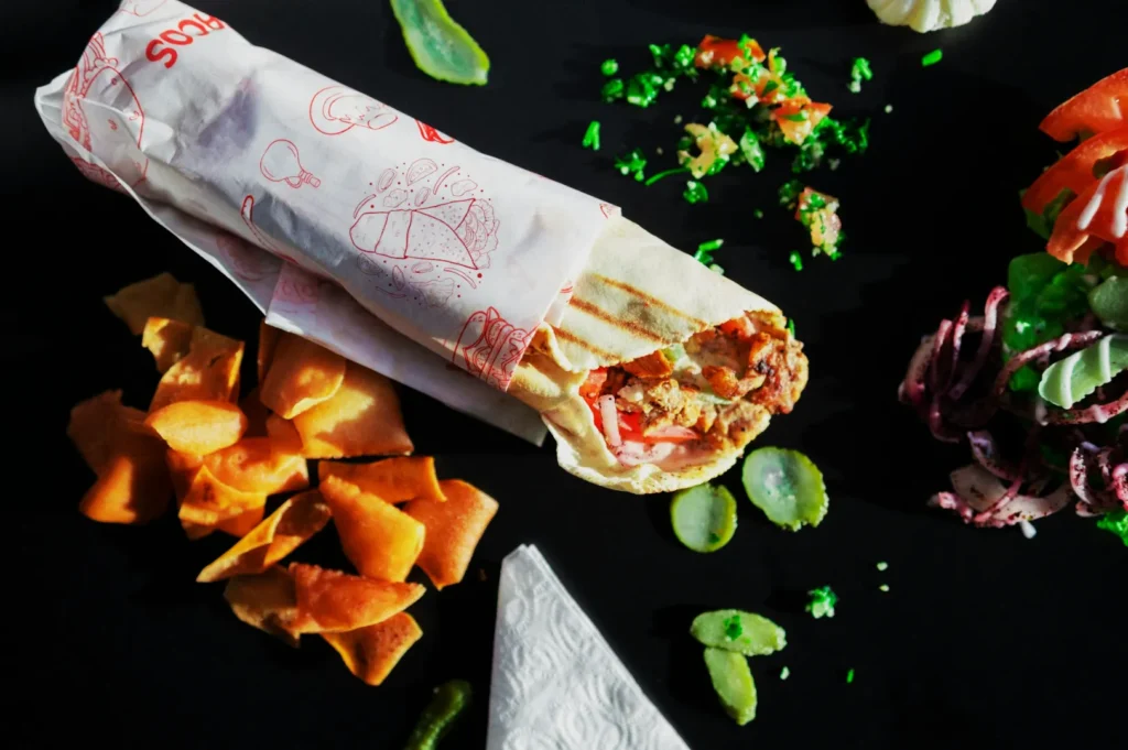 A wrapped shawarma on a black surface surrounded by vibrant toppings: sliced vegetables, herbs, pickles, chips, and a napkin. Bright and appetizing scene.