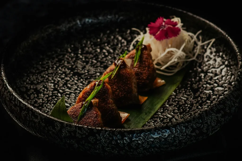 Plated dumplings on a dark textured dish, garnished with sauce and green herbs. A nest of radish shreds and a pink flower add elegance.