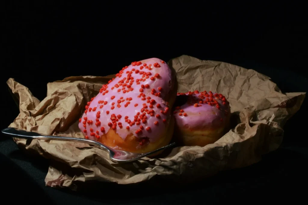 Two heart-shaped donuts with pink icing and red sprinkles rest on crumpled paper, beside a metal spoon. The background is dark, creating a dramatic contrast.