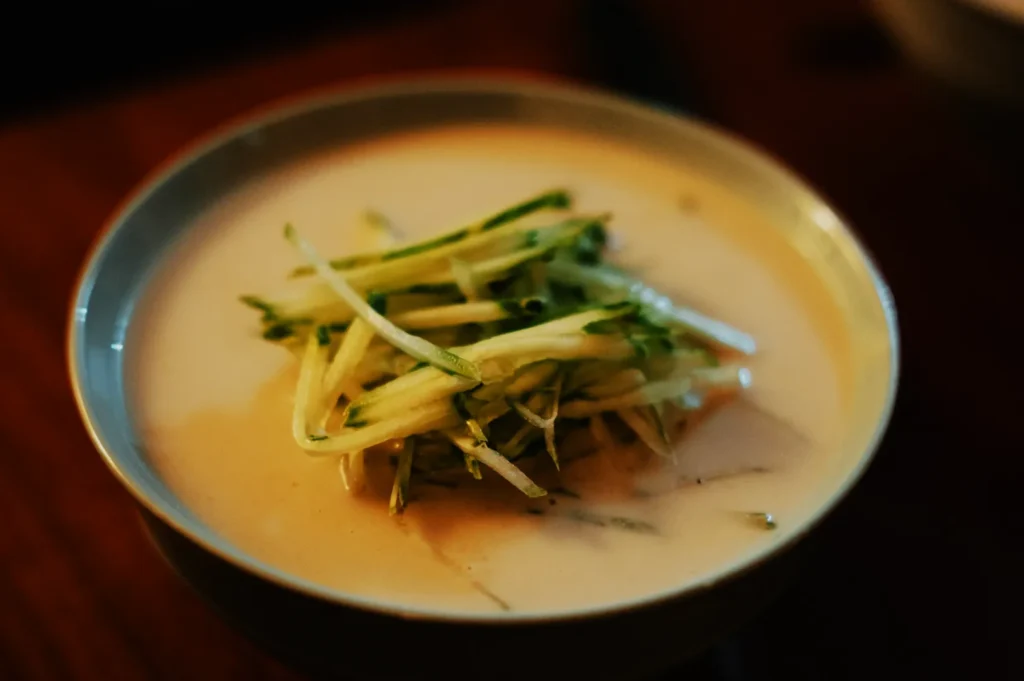 A bowl of creamy, light soup topped with thinly sliced, fresh cucumber strips. The setting is warm and inviting, evoking a sense of comfort.