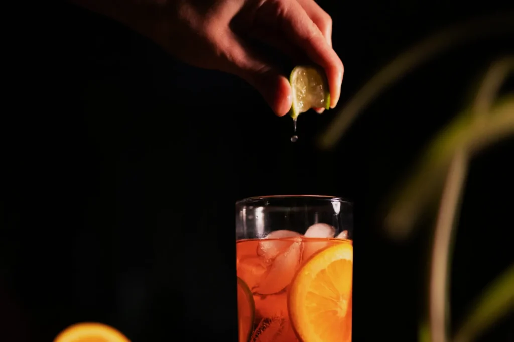 A hand squeezes a lime over a glass filled with iced tea, garnished with orange slices. The image conveys freshness and a refreshing summer vibe.