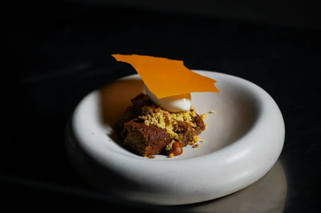 A small, artfully plated dessert on a round white dish. It features crumbled cake, a dollop of cream, caramel drizzle, and an orange garnish.