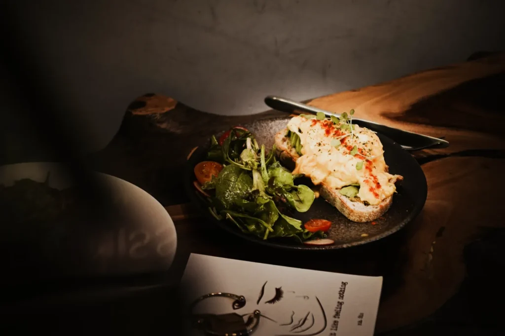 A rustic wooden table holds a plate with an open-faced sandwich topped with scrambled eggs and herbs, accompanied by a fresh salad with cherry tomatoes.