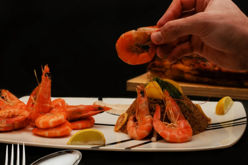 A hand is picking up a shrimp from a platter of cooked shrimp and rice garnished with lemon wedges and dill, set against a dark background.