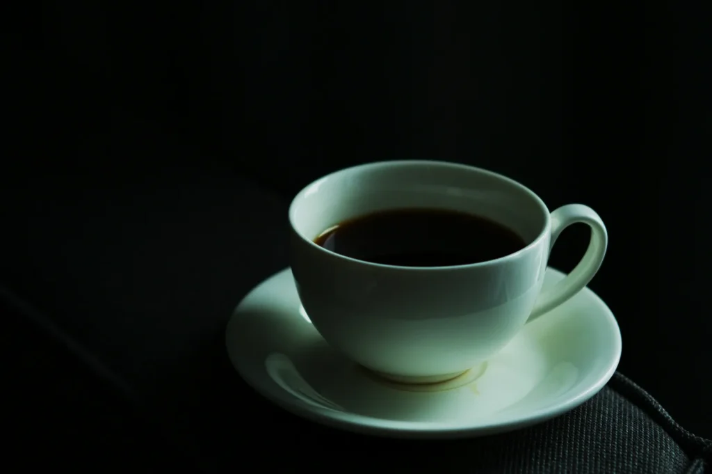 A white cup filled with black coffee sits on a matching saucer against a dimly lit, dark background, exuding a calm and reflective atmosphere.