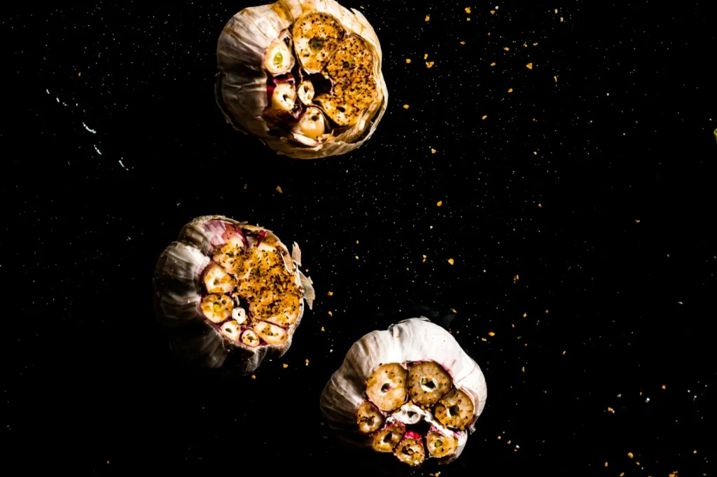 Three roasted garlic bulbs on a black surface, sprinkled with seasoning, creating a rustic and earthy feel with scattered spices around.