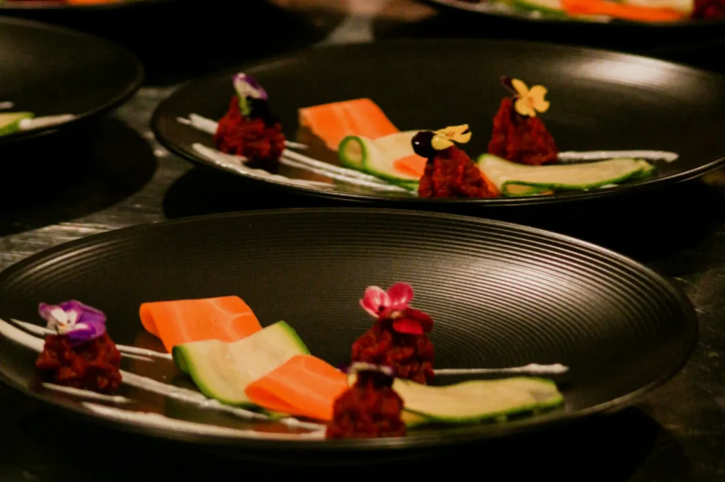 Elegantly plated gourmet dish with vibrant vegetables, garnished with edible flowers and delicate sauces on black plates, conveying a sophisticated tone.