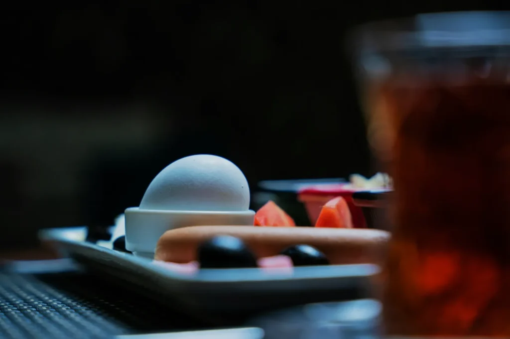 A close-up of a white egg, various fruits, and a sausage on a white plate. The focus is on the egg, with a blurred drink in the foreground.
