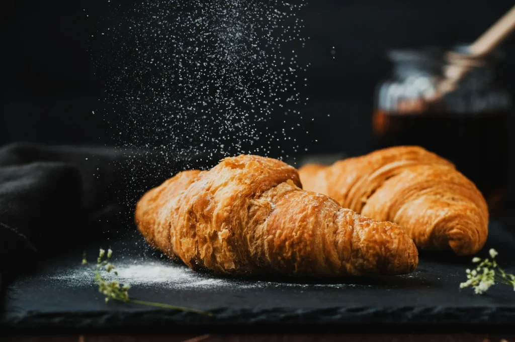 Golden croissants on a slate board are dusted with powdered sugar against a dark background, next to a jar of honey, creating a warm, rustic atmosphere.