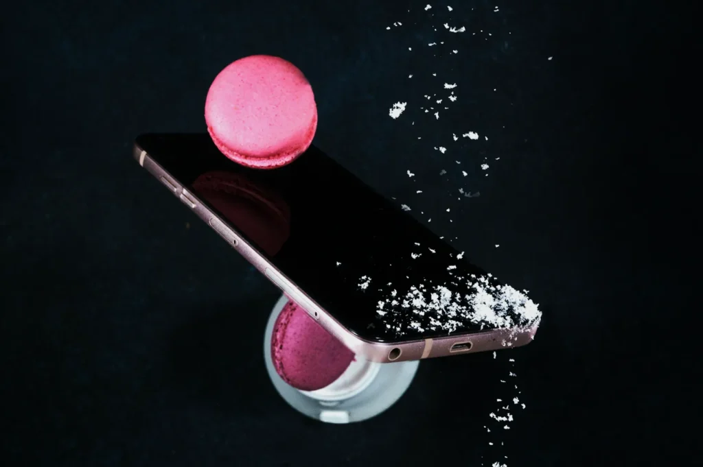 A pink macaron balanced on a smartphone, with another underneath on a stand. Powdered sugar sprinkles create a whimsical, artistic scene on a black background.