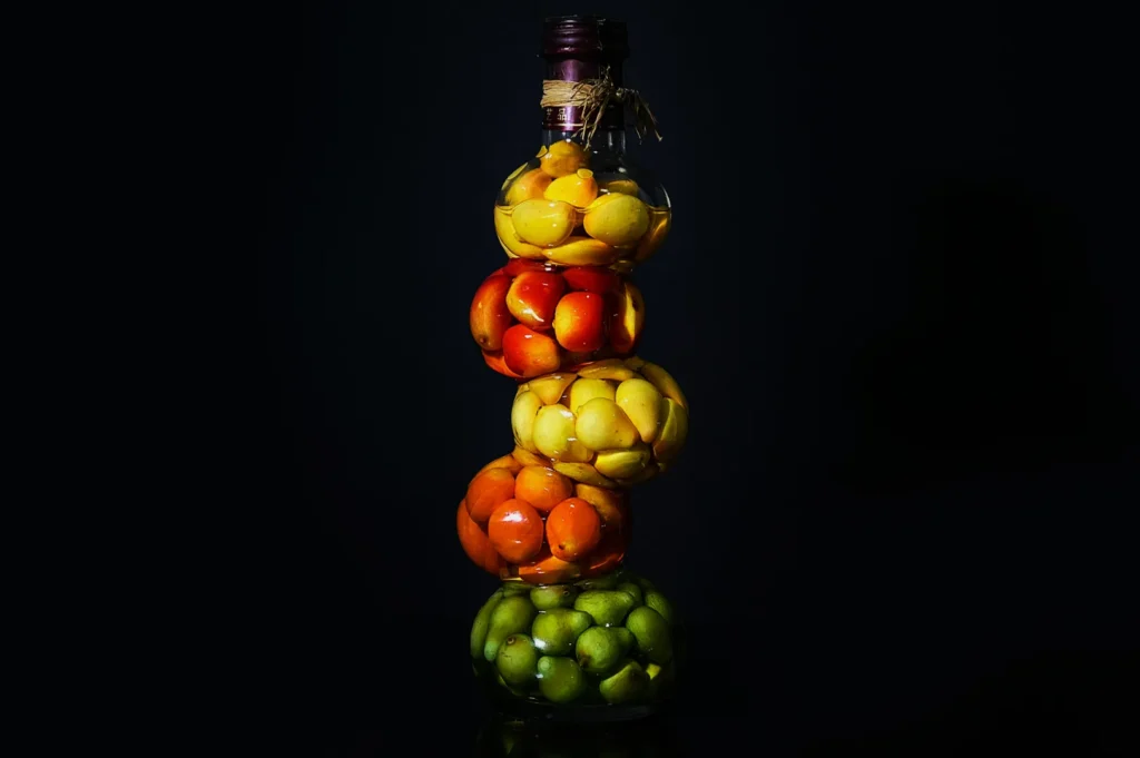 A tall, clear bottle segmented into four sections, each filled with colorful pickled fruits: green, orange, red, and yellow, set against a dark background.