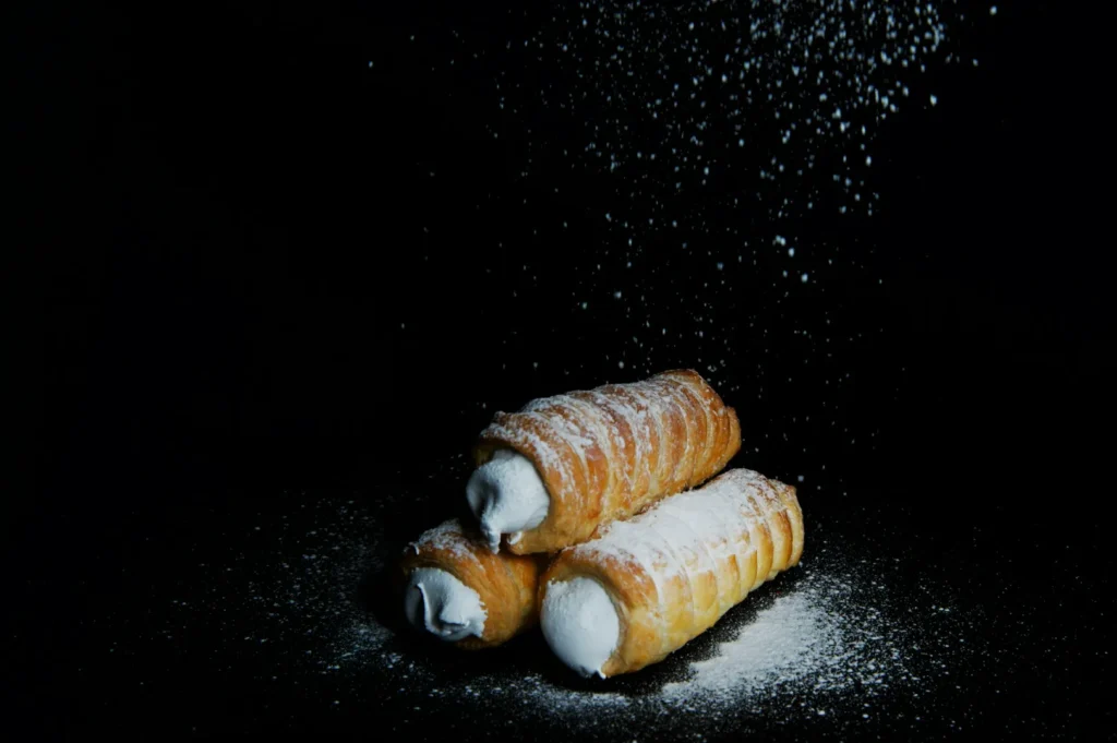 Three cream-filled pastries stacked on a dark background, dusted with powdered sugar falling from above, creating a delectable and inviting scene.
