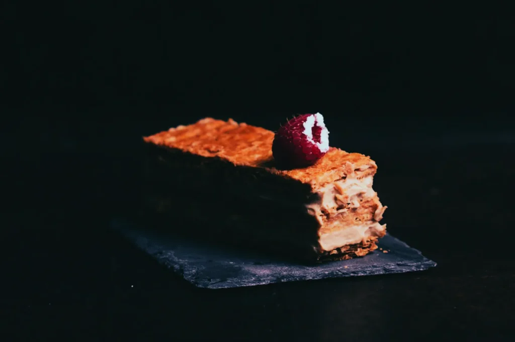 A piece of layered puff pastry dessert topped with a red raspberry on a dark slate. The dessert's textures are highlighted against a dark backdrop.