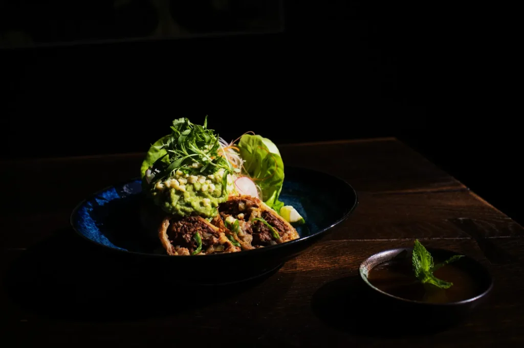 A blue bowl with a halved wrap filled with beef and topped with avocado and greens sits on a dark wooden table, creating a moody, elegant ambiance.