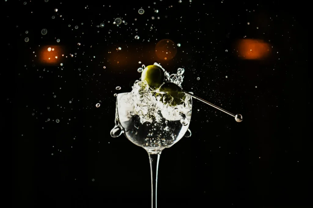 A martini glass with liquid splashing out, pierced olives on a toothpick inside, set against a dark background with soft bokeh lights. Elegant and dynamic.