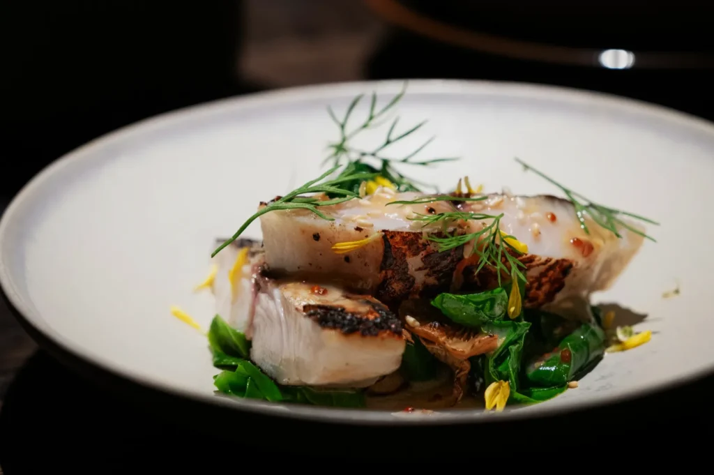 Grilled fish on a white plate garnished with fresh herbs, atop greens and mushrooms. The dish is artfully arranged, with a sophisticated ambiance.